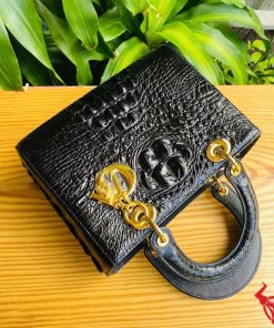 Hình ảnh túi xách TDA4 màu đen, da cá sấu gù cao cấp, khóa vàng nổi bật, đặt trên bàn gỗ với cây xanh và mô tô.