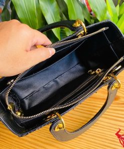 Hình ảnh túi xách TDA4 màu đen, da cá sấu gù sang trọng, khóa vàng lấp lánh, đặt trên bàn gỗ với cây xanh phía sau.