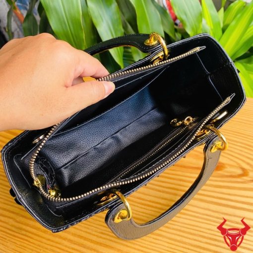 Hình ảnh túi xách TDA4 màu đen, da cá sấu gù sang trọng, khóa vàng lấp lánh, đặt trên bàn gỗ với cây xanh phía sau.