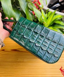 Hình ảnh túi xách TDA4 màu xanh lá, da cá sấu cao cấp, khóa vàng nổi bật, đặt trên bàn gỗ với cây xanh phía sau.