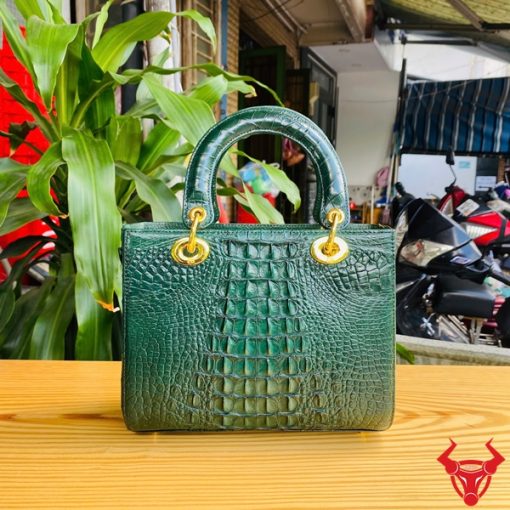 Hình ảnh túi xách TDA4 màu xanh lá, da cá sấu sang trọng, khóa vàng lấp lánh, đặt trên bàn gỗ với cây xanh và mô tô.