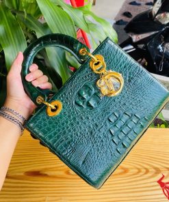 Hình ảnh túi xách TDA4 màu xanh lá, da cá sấu cao cấp, khóa vàng lộng lẫy, đặt trên bàn gỗ với nền cây xanh.