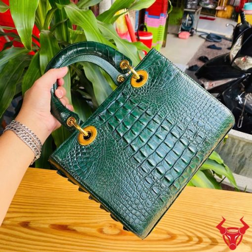 Hình ảnh túi xách TDA4 màu xanh lá, da cá sấu cao cấp, khóa vàng nổi bật, đặt trên bàn gỗ với cây xanh và mô tô.