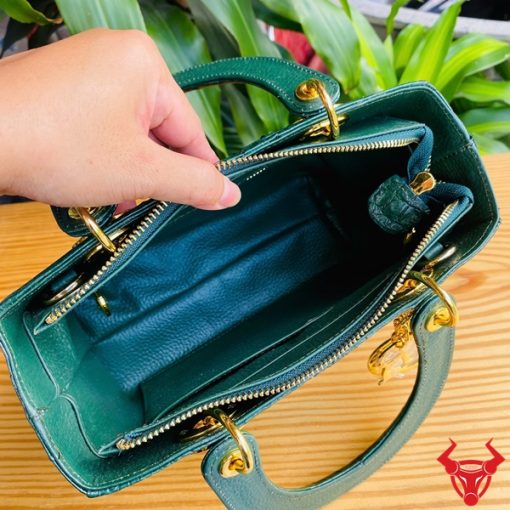 Hình ảnh túi xách TDA4 màu xanh lá, da cá sấu sang trọng, khóa vàng lấp lánh, đặt trên bàn gỗ với cây xanh phía sau.
