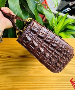Hình ảnh túi xách TDA4 màu nâu đất, da cá sấu cao cấp, khóa vàng nổi bật, đặt trên bàn gỗ với cây xanh phía sau.
