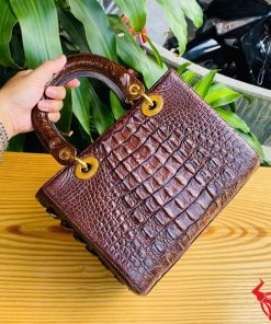 Hình ảnh túi xách TDA4 màu nâu đất, da cá sấu sang trọng, khóa vàng lấp lánh, đặt trên bàn gỗ với cây xanh và mô tô.