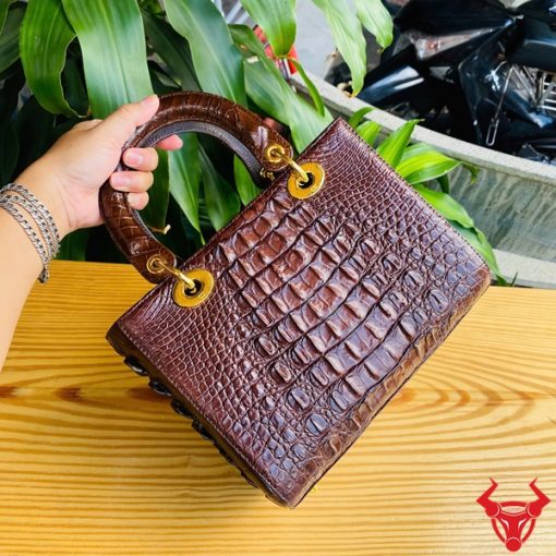 Hình ảnh túi xách TDA4 màu nâu đất, da cá sấu sang trọng, khóa vàng lấp lánh, đặt trên bàn gỗ với cây xanh và mô tô.