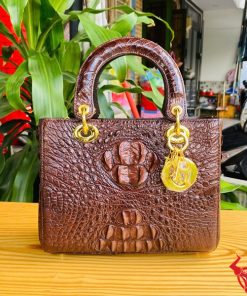 Hình ảnh túi xách nữ da cá sấu TDA4 màu nâu đất, thiết kế sang trọng, khóa vàng lấp lánh, đặt trên bàn gỗ với cây xanh và mô tô.