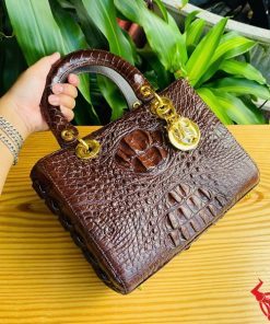 Hình ảnh túi xách TDA4 màu nâu đất, da cá sấu cao cấp, khóa vàng lộng lẫy, đặt trên bàn gỗ với nền cây xanh.