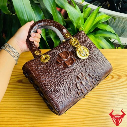 Hình ảnh túi xách TDA4 màu nâu đất, da cá sấu cao cấp, khóa vàng lộng lẫy, đặt trên bàn gỗ với nền cây xanh.