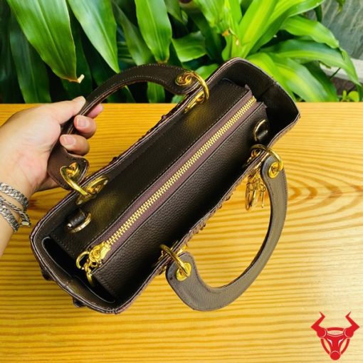 Hình ảnh túi xách TDA4 màu nâu đất, da cá sấu cao cấp, khóa vàng nổi bật, đặt trên bàn gỗ với cây xanh và mô tô.