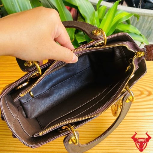 Hình ảnh túi xách TDA4 màu nâu đất, da cá sấu sang trọng, khóa vàng lấp lánh, đặt trên bàn gỗ với cây xanh phía sau.