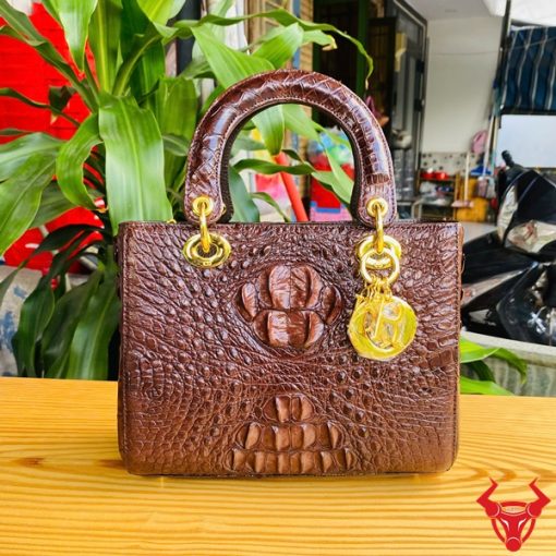 Hình ảnh túi xách nữ da cá sấu TDA4 màu nâu đất, thiết kế sang trọng, khóa vàng lấp lánh, đặt trên bàn gỗ với cây xanh và mô tô.