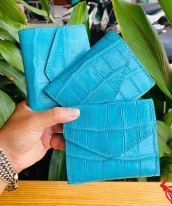 Ảnh cầm ba ví nữ mini da cá sấu VRAA8 màu turquoise, thiết kế 3 gấp, nhấn mạnh thời trang, phong cách nhỏ gọn trên nền thực vật xanh.