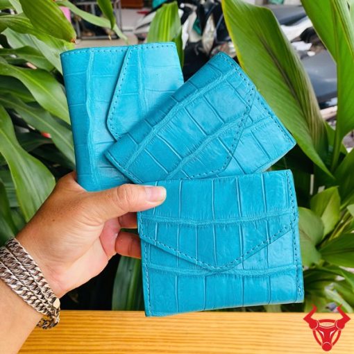 Ảnh cầm ba ví nữ mini da cá sấu VRAA8 màu turquoise, thiết kế 3 gấp, nhấn mạnh thời trang, phong cách nhỏ gọn trên nền thực vật xanh.