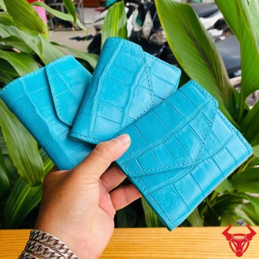 Ba chiếc ví nữ 3 gấp mini da bụng cá sấu VRAA8 xanh dương trong tay, nền lá cây, đại diện cho sự thời trang, phong cách và nhỏ gọn.