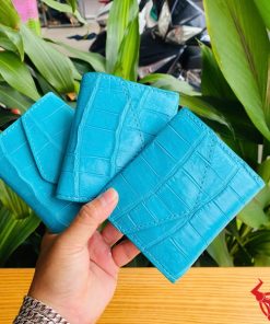 Hình ảnh ba chiếc ví nữ 3 gấp mini da bụng cá sấu VRAA8 màu xanh dương được cầm trên tay, nền xanh lá cây, thể hiện sự thời trang, phong cách và nhỏ gọn.