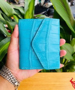 Ảnh ba ví nữ mini VRAA8 da bụng cá sấu xanh dương được cầm, nền lá cây, nhấn mạnh thời trang, phong cách và thiết kế nhỏ gọn.
