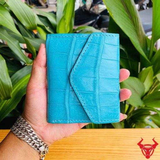 Ảnh ba ví nữ mini VRAA8 da bụng cá sấu xanh dương được cầm, nền lá cây, nhấn mạnh thời trang, phong cách và thiết kế nhỏ gọn.