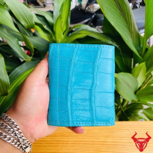 Hình ảnh cầm ba ví nữ VRAA8 da bụng cá sấu 3 gấp mini xanh dương, nền thực vật, thể hiện thời trang, phong cách và nhỏ gọn.