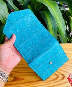 Ảnh ba ví mini 3 gấp da cá sấu VRAA8 màu xanh cầm tay, nền lá cây xanh, nhấn mạnh thời trang, phong cách nhỏ gọn cho sản phẩm.