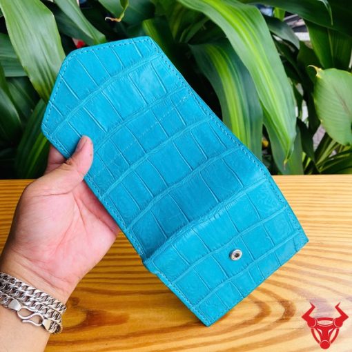 Ảnh ba ví mini 3 gấp da cá sấu VRAA8 màu xanh cầm tay, nền lá cây xanh, nhấn mạnh thời trang, phong cách nhỏ gọn cho sản phẩm.